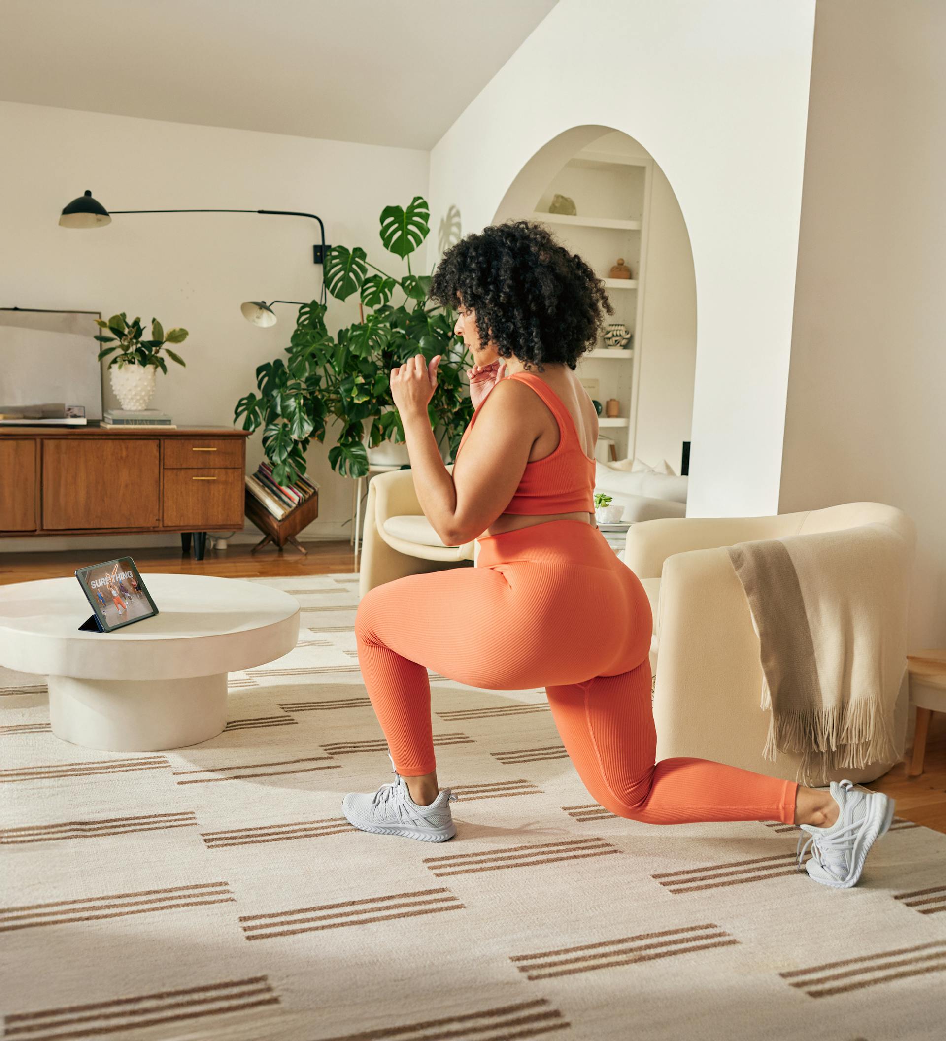 A woman in workout clothes doing lunges in front of a tablet in her livingroom