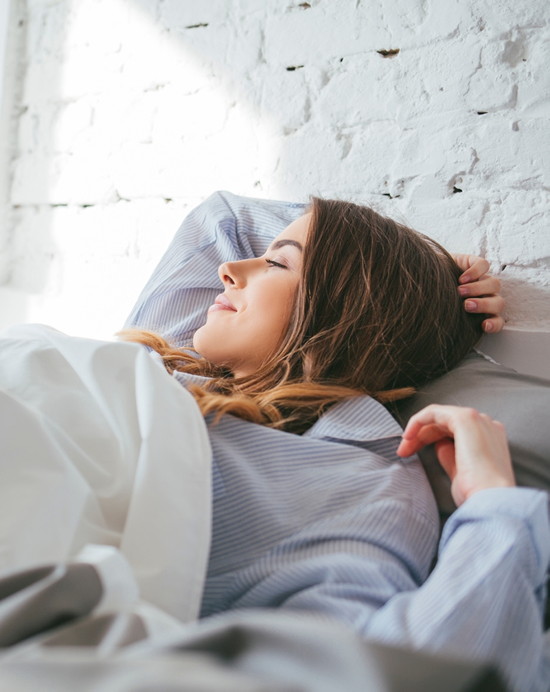 Young woman lying in bed, smiling towards her sun-filled window with a hand over her head