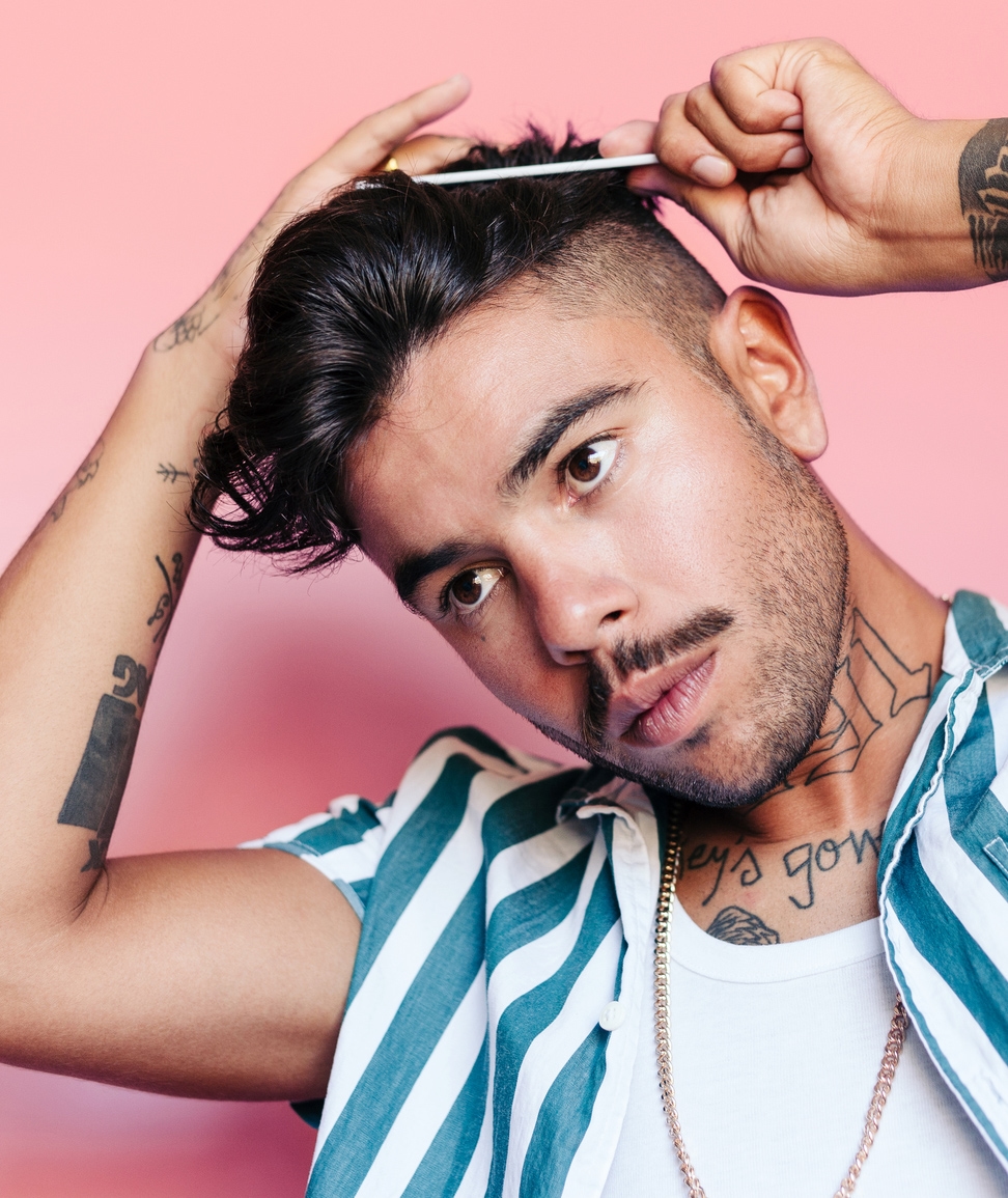 Young, hip latino man with slick black hair and tattoos wearing jean shirt combing his hair and tilting his head