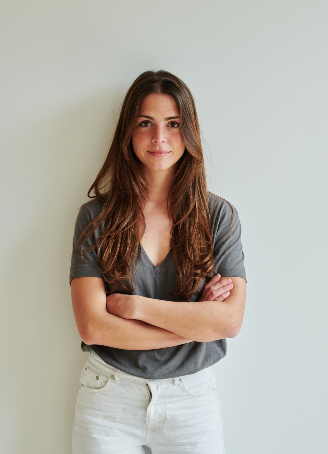 Young white woman standing against wall with arms crossed looking intently at camera