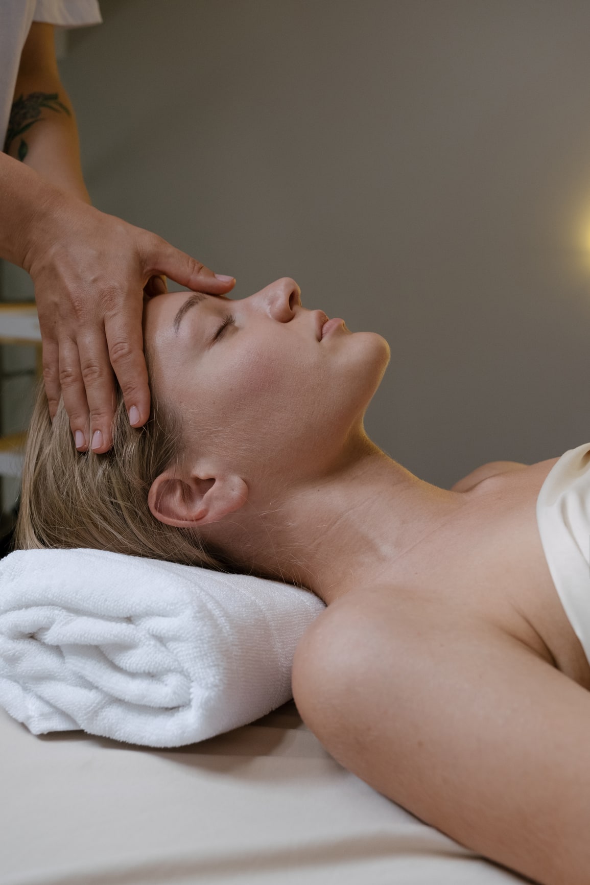 A woman lying on a massage table with eyes closed, receiving a forehead massage from a person standing beside her.
