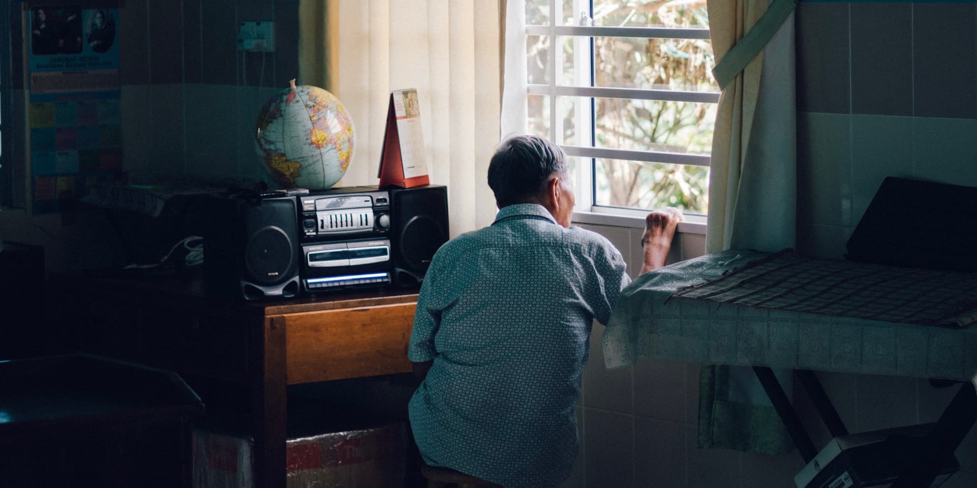 Fotografía de un hombre de espaldas a la cámara, sentado solo y mirando por la ventana en una habitación con poca luz.