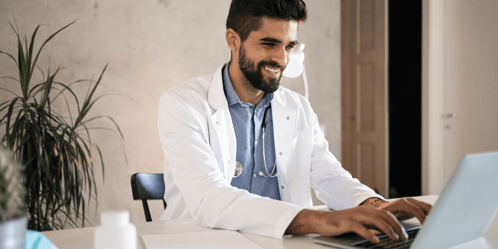 A male doctor with medium-toned skin and dark hair sits in a blue shirt and lab coat with a stethoscope around his neck, typing into a computer and smiling