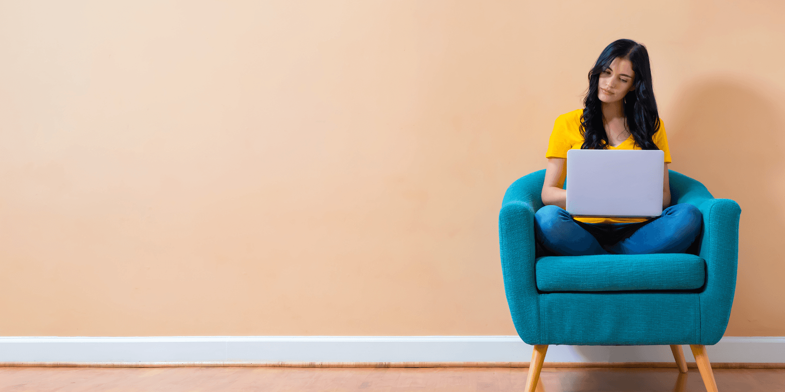 A young woman with long dark hair wearing a yellow t-shirt sits on a blue velvet chair against a pink-tinged wall, typing into a laptop.