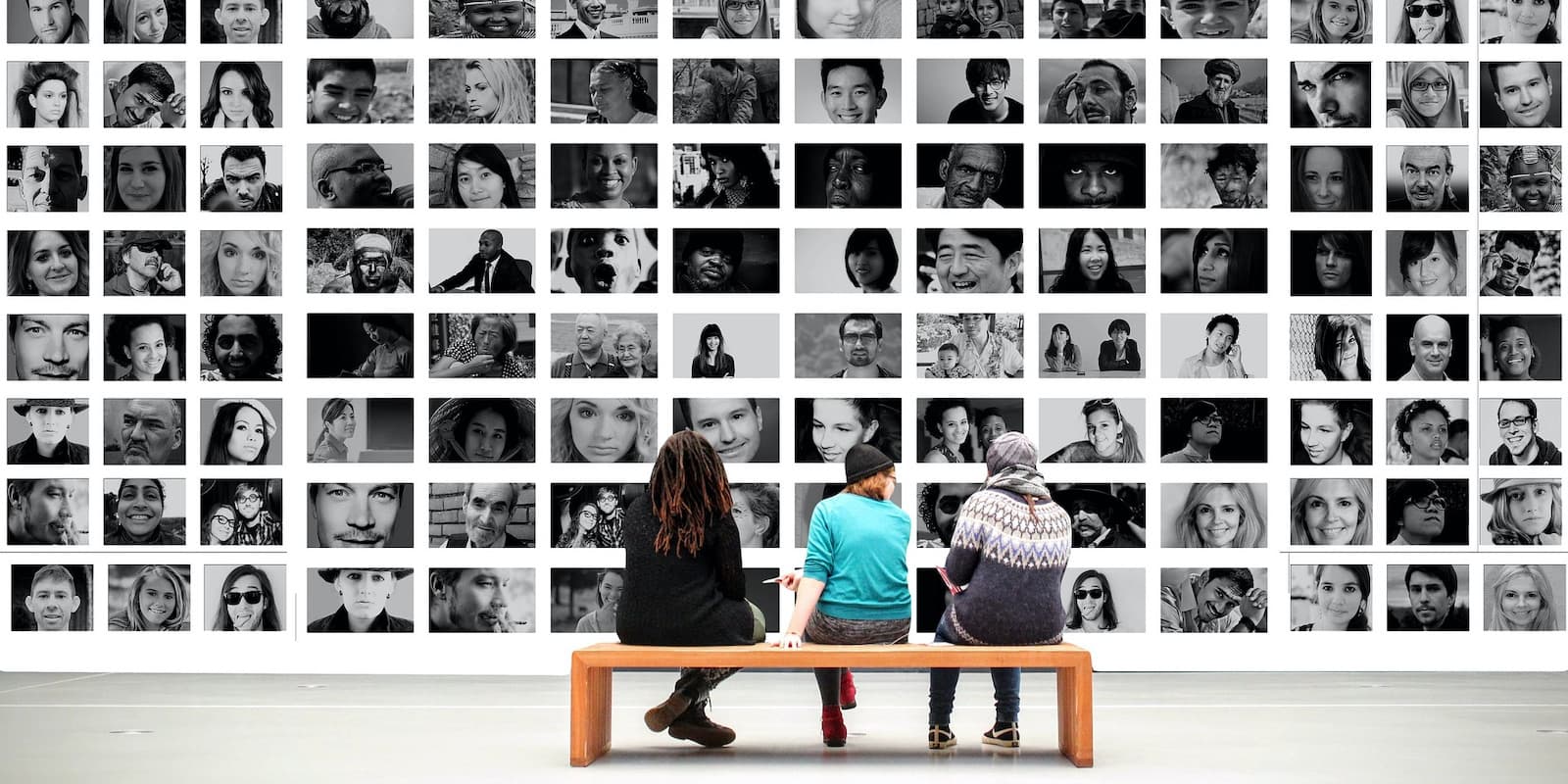 Three young people sit with their backs to the camera in front of a large museum exhibit of dozens of black-and-white photographs.