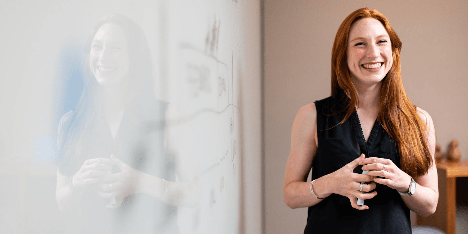 A young woman with long red hair wearing a black tank top smiles into the camera as she stands next to an office whiteboard.