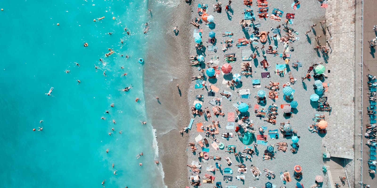 Una fotografía en lo alto de lo alto de una playa mediterránea con aguas turquesas a la izquierda y arena salpicada de sombrillas y personas a la derecha.