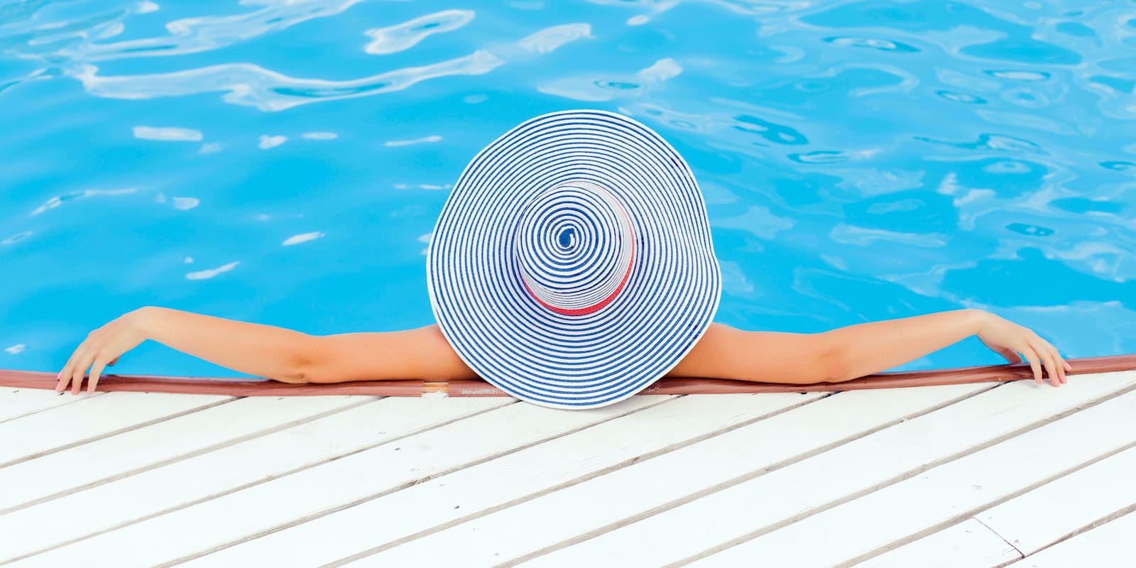 A photograph of a woman in a pool looking away from the camera. She's wearing a large-brimmed hat and has her arms stretched out alongside her.