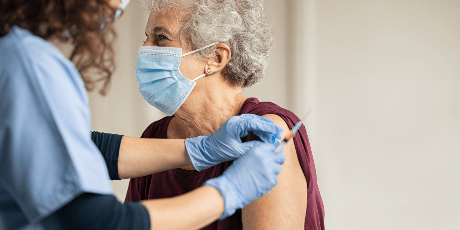 A white senior woman with short gray hair wearing a medical mask and red t-shirt is given an injection on her upper arm by a medical technician with curly hair wearing a dark long-sleeved shirt and scrubs.