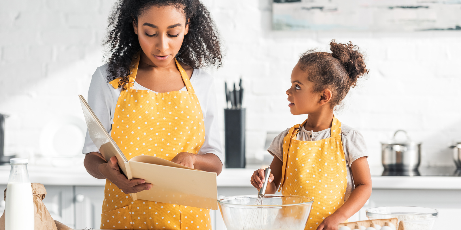 Two Black sisters wearing white tshirts and yellow polka dotted aprons bake gluten free treats in a modern white kitchen, with the addorable toddler sister looking up adoringly at her big sister who is reading from a cookbook.