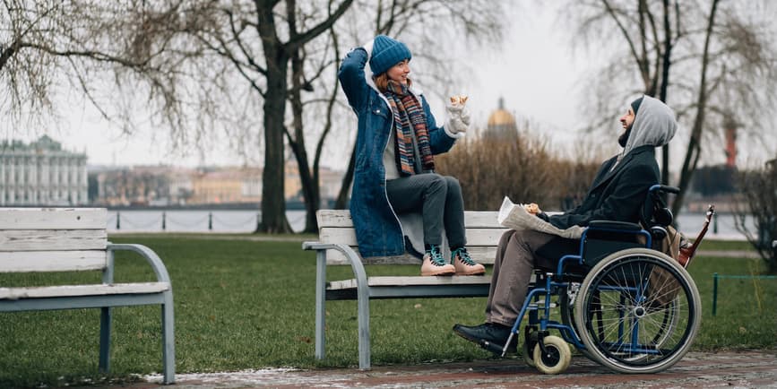 A color photograph of a wide outdoor winter landscape, with two friends in a park with water and bare trees behind them. A young white woman sits on the top ledge of a park bench, wearing jeans, a scarf, a winter coat and hat, smiling down at her friend. A white man in a wheelchair wearing khaki pants, a dark jacket and sweatshirt with the hood up laughs up to her.