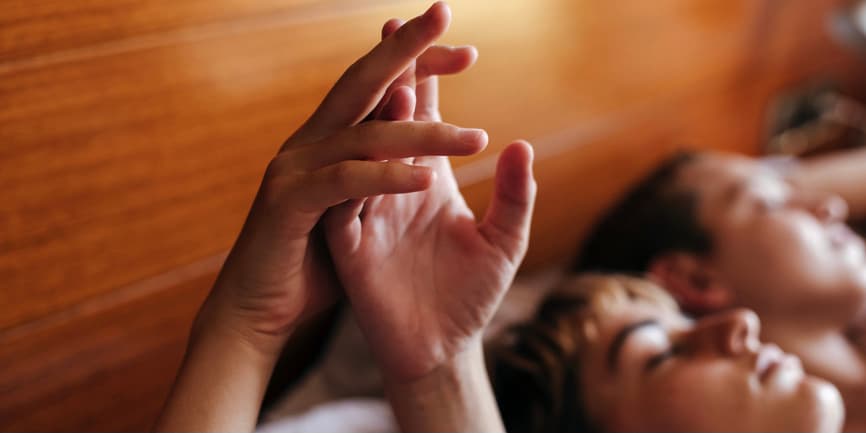 A color photograph of two white people laying in bed against a wooden headboard, their hands intertwined in the air.