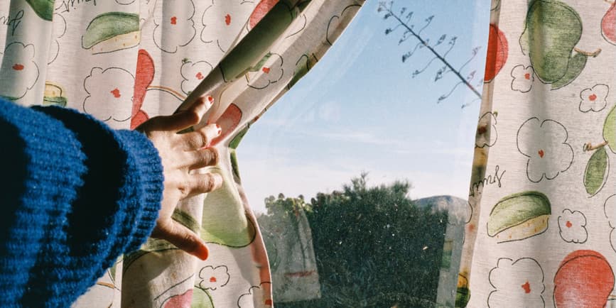 Una fotografía en color de una ventana con un brazo en un suéter azul tirando hacia atrás cortinas descoloridas con frutas, un cielo azul que muestra el exterior.