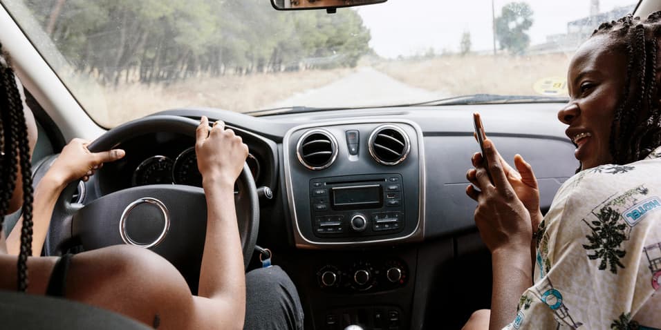 A color photograph from the backseat of a car looking into the front, where two Black women are driving, smiling and taking photographs.