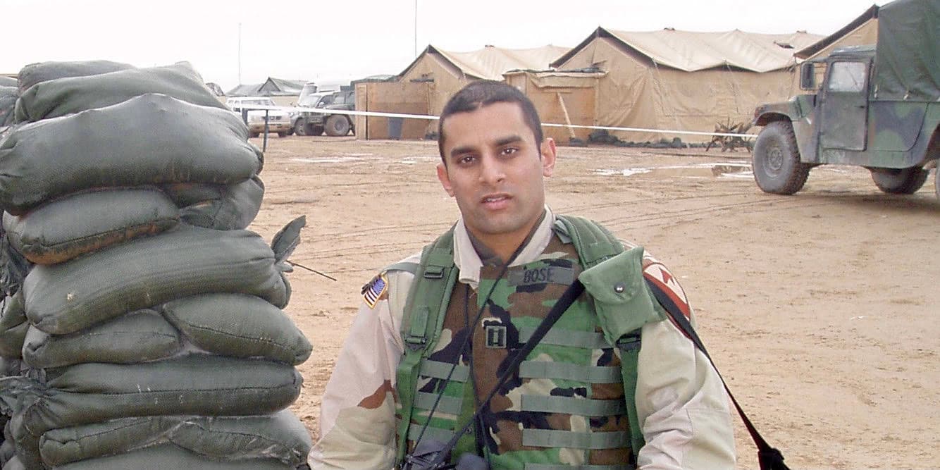 A photograph of Dr. Sudip Bose in his military fatigues, standing in front of a line of Army tents and a military truck.