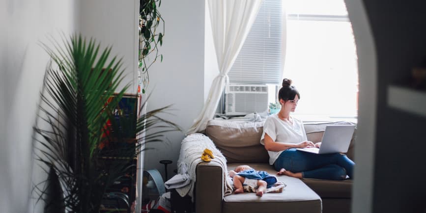 A color photograph of a white woman with her long hair tied up, working on a computer while seated in her living room with a baby resting on the couch next to her.