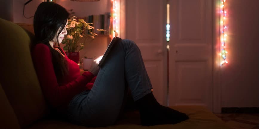 A color indoor photograph of young woman sitting on their bed, wearing jeans and a red long-sleeve shirt, shopping on their iPad