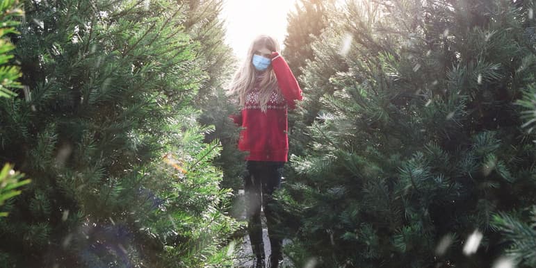 A color outdoor photograph of a young girl wearing jeans, a red sweater and medical face mask, standing between uncut Christmas trees.