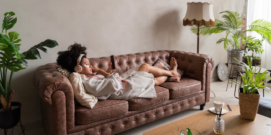 A color indoor photograph of young Black woman with an afro haircut laying on a brown couch, surrounded by a few plants. She's wearing a short silky robe, headphones and a face mask and looking at her phone.