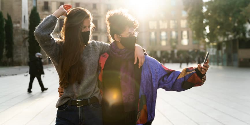 A color outdoor photograph of a young white woman with long brown hair and a young Black woman with her black hair tied up. They're wearing long sweaters and black N95 masks, cuddling together as they take a selfie.