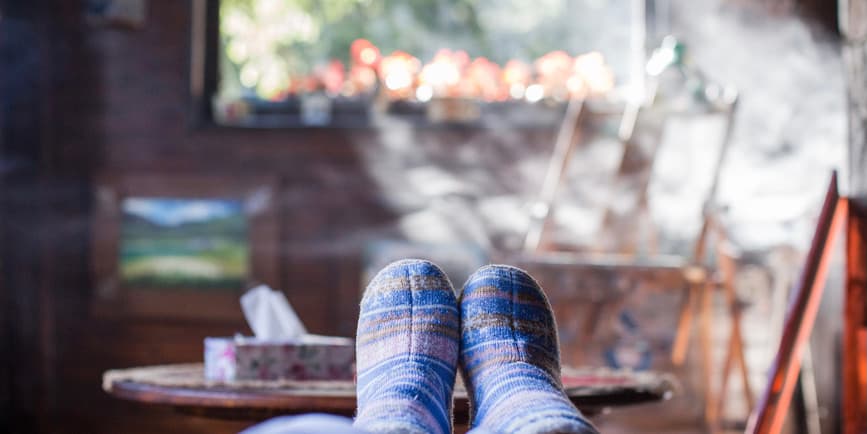 A color indoor photograph of a cheerful winter scene, with slippered feet perched on a coffee table in front of a misty bright window with holiday lights outdoors.