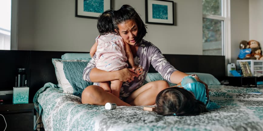 A color indoor photograph of a young Asian woman and her two daughters on a bed with blue and white pillows and such around them. The mother has a toddler perched over her should while she leans down over the other, who is laying on the bed.