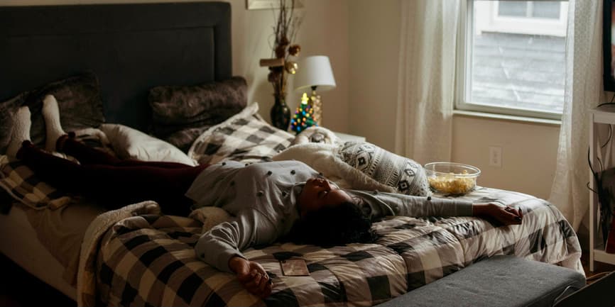 A color indoor photograph of a white woman wearing pajamas and lying on her bed face up, a bowl of popcorn nearby and a small ceramic Christmas tree glowing from the nightstand.