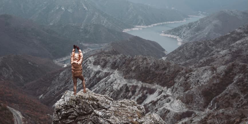 A color outdoor landscape of a mountain range with a river cutting through it. In the foreground a woman stands with her back to the camera, wearing brown pants and a shiny gold jacket, raising their arms to the sky.