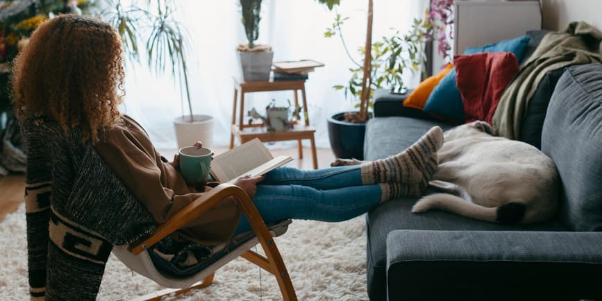 A color indoor photograph of a young Black woman wearing jeans and a sweater, her full brown hair hiding her face, reading a book with her legs and socks perched on a blue couch where a dog lays sleeping.