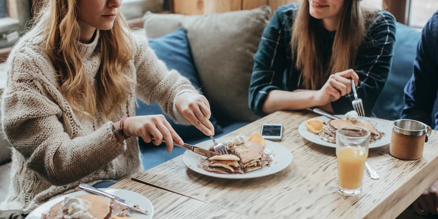 A color indoor photograph of two white women with long hair wearing sweaters. They sit at a restaurant table, cutting into pancakes.