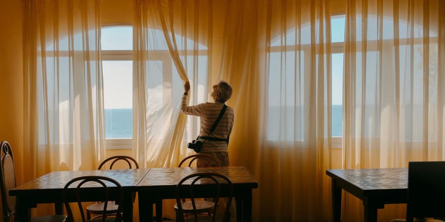 A color indoor photograph of a white, older male tourist wearing a camera around their neck, standing next to a window amongst bare dining tables, opening gauzy yellow curtains to a view of water and sky.