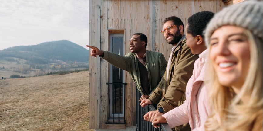 A color outdoor photograph of a group of friends on a deck against a wood home, with mountains in the distance. They're smiling off the camera, with one pointing to the distance.