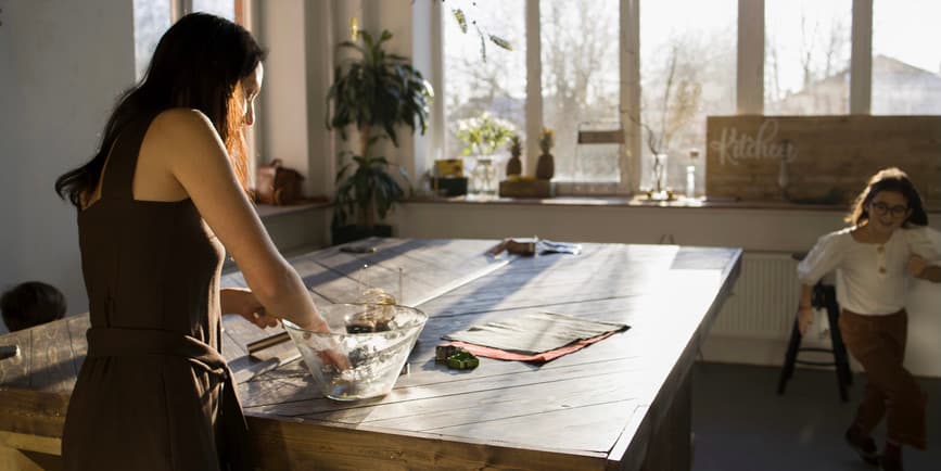 A color photograph of a sun-dappled kitchen, where a white mother with long dark hair wearing an apron kneads bread dough as her toddler runs around her.