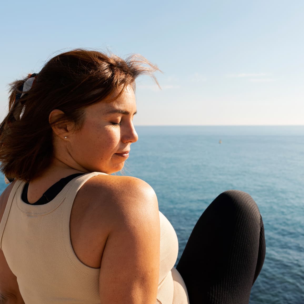 Woman sitting in front of the ocean, eyes closed, head turned toward her shoulder and the sunshine.