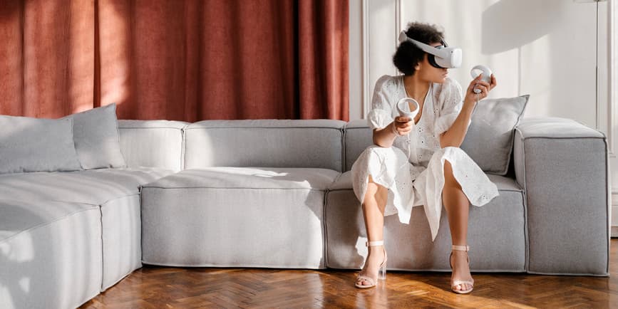 A color indoor photograph of a young Black woman with short brown hair wearing a long white dress, sitting on a gray couch in front of a dark red curtain, wearing virtual reality goggles and looking off camera.