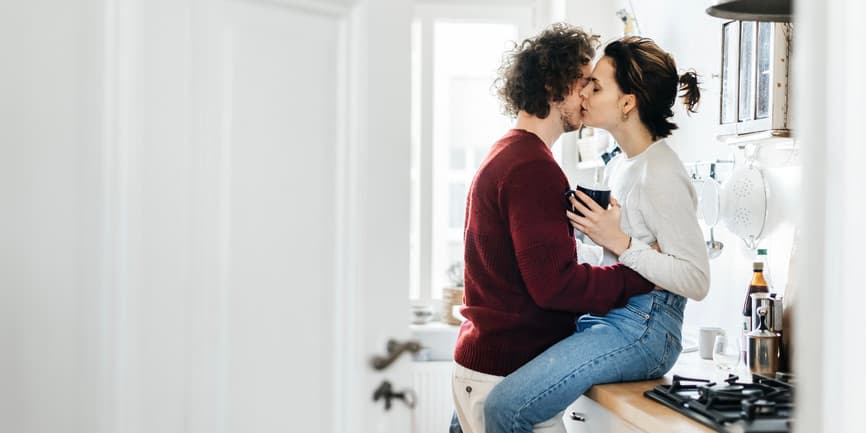 An indoor color photograph of a white kitchen, where a young white man in a dark red sweater embraces a young white woman with tied up brown hair, a white shirt and jeans who's sitting on the counter holding a coffee cup.