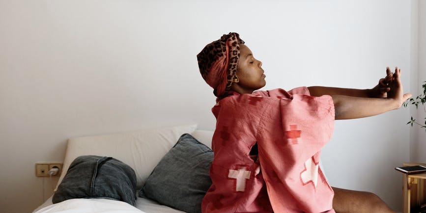 Fotografía en color de una mujer negra en su dormitorio, estirándose mientras está sentada en su cama. Lleva un camisón rosa-rojo y una diadema.