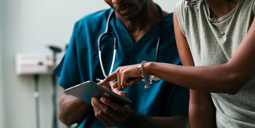 A closeup color photograph of a Black woman wearing a sleeveless top points at a screen a young Black doctor wearing blue scrubs is holding.