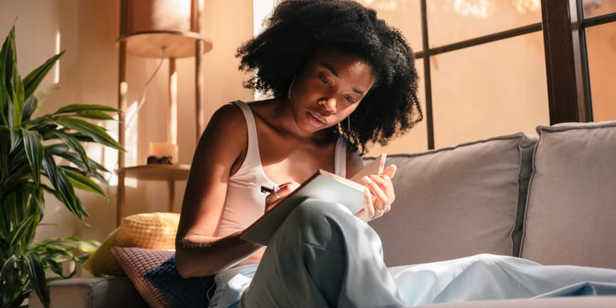 An indoor color photograph of a young Black teen with Black hear wearing a pink tank and pajama pants, sitting on a catch in a room with pink walls and a plant, writing in a journal on her lap.