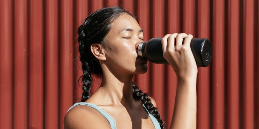 Una mujer de pelo largo y negro trenzado lleva puesta una camiseta deportiva mientras bebe de una botella de agua con los ojos cerrados
