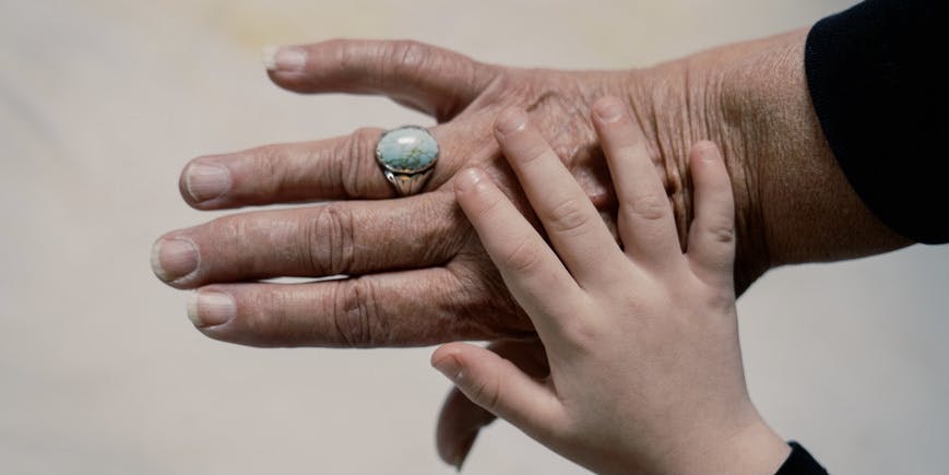 Fotografía en color de la mano de un niño apoyada sobre la mano de una persona mayor, con la piel ligeramente arrugada, que lleva un anillo turquesa.