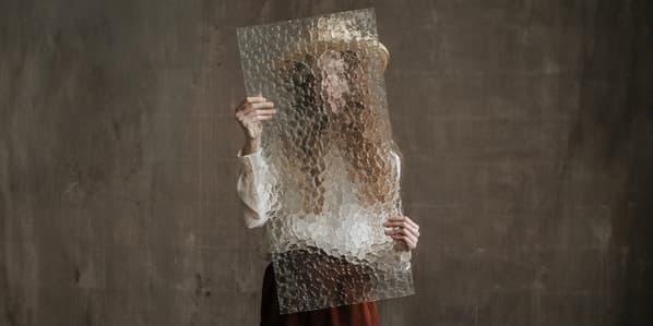 A color studio portrait of a thin white woman with long blond hair wearing a brown skirt, white blouse and tan cowboy hat, standing against a brown wall and holding a textured piece of glass, which obscures her face.