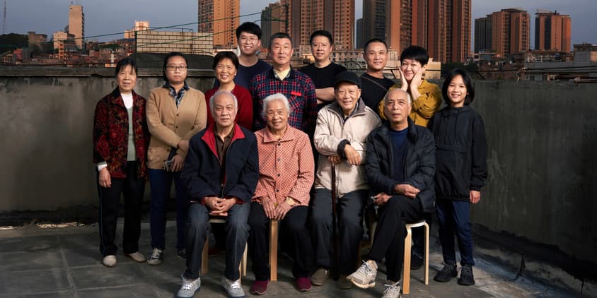 A color outdoor photograph of an Asian family standing on a rooftop against a sunset city skyline. They are more than ten people crossing generations, from children to grandparents.