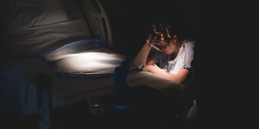 A color interior photograph of a dark hospital room where a white woman with short curly hair wearing a hospital gown kneels down next to the bed, where a light shines down on it.