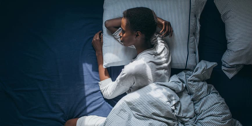 A thin Black woman with short black hair wearing white pajamas lays sleeping on a striped blue and white pillow and blue sheets.