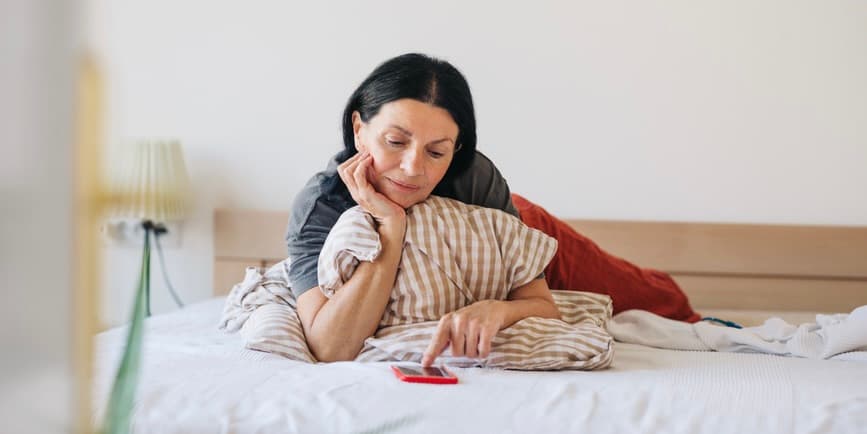 A middle aged woman with dark hair is laying in bed looking at a phone in a red case