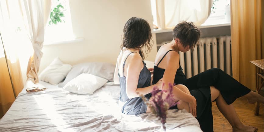 A color photograph of two white women in pajamas sitting on the edge of a bed in a sunny room, both with their heads down and one sitting behind the other, rubbing their back