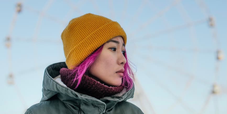 Retrato de una joven asiática de pelo rosa con una gorra de invierno amarilla, un abrigo grueso y un suéter de cuello alto, mirando fuera de cámara con el London Eye de fondo.