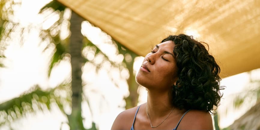 An outdoor photograph of a beautiful brown-skinned woman with black curly hair. She's sitting underneath an umbrella with palm trees in the background, her eyes shut and face tilted up to the sky.