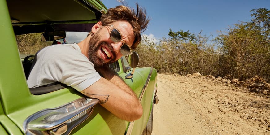 A color outdoor photograph of a young white man with brown hair and a beard wearing a white t-shirt, leaning out of a green vintage car window and smiling into the camera.